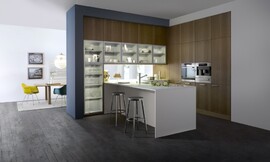 Een harmonieuze drieklank van materialen: keukenfronten met natuurlijk werkend houten structuuroppervlakte, matte magnolia kunststof en een dun wit aanrechtblad/keukenblok. Glazen deurtjes voor de servieskast maken het geheel af. Zuordnung: Stil Klassieke keukens, Planungsart L-vormige keuken