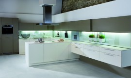 Deze keuken in F-vorm (!) valt op door het contrast tussen gebroken witte onderkasten, brons-bruine bovenkasten en een verlichte glazen keukenachterwand. Een gedeelte van de keukenwand zweeft boven de grond. Boven het centrale kookgedeelte hangt een opvallende afzuigkap. De keuken heeft een dun aanrechtblad dat optisch niet opvalt. Zuordnung: Stil Design-keukens, Planungsart keukenblok