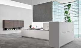Strakke lijnen zonder onnodige versierselen, helder wit en een moderne uitstraling zijn de kenmerken van deze keuken in eilandvorm. Het rechte keukenblok bestaat uit onderkasten met deurtjes en grote lades die ideaal zijn voor het opbergen van serviesgoed, potten en pannen. De rechterhoek van de keuken wordt omsloten door half hoge kasten. Op de achtergrond is een zeer bijzonder glazen open kastsysteem te zien dat aan het plafond is opgehangen. Zuordnung: Stil Design-keukens, Planungsart Detail keukenontwerp