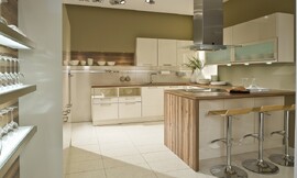 In deze U-vormige keuken is gebruik gemaakt van frisse witte fronten gecombineerd met een houten aanrechtblad dat aan de zijkant van het kookeiland doorloopt en ook als achtergrond bij een legplank is gebruikt. Aan de linkerkant zijn twee onderkasten opgenomen waarvan het bovenste gedeelte open is. Daarnaast komt de spoelkast en nog een onderkast. Boven de spoelbak zijn drie hangkastjes opgenomen. Links van de hangkastjes is een legplank bevestigd tegen hetzelfde hout als het aanrechtblad. Het andere wandgedeelte bestaat uit onderkasten, handige werkruimte op het aanrechtblad met daarboven twee brede kasten met glazen inzet en daaronder spotjes. Haaks hierop staat het kookeiland met fornuis, afzuigkap en aan de andere kant een gezellige zitgelegenheid. In de hoge wandkasten is de apparatuur opgenomen. Zuordnung: Stil Moderne keukens, Planungsart U-vormige keuken