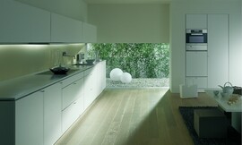 Deze zeer kalm ogende keuken heeft mat witte fronten met subtiele grepen. Een kast is ingebouwd in de muur, voor een optisch rustig effect en de roestvrijstalen oven is op handige ooghoogte in deze kast aangebracht. Het brede keukenblok met verlichting in de bovenkasten staat op een sokkel en heeft een dun aanrechtblad in dezelfde tint.De greeploze bovenkasten zijn vrij breed en niet zo hoog. Onder één van de bovenkasten is de afzuigkap vrijwel onzichtbaar bevestigd. Zuordnung: Stil Design-keukens, Planungsart keukenblok