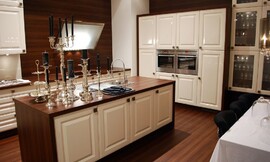 Aanrechtbladen van donker hout en mat witte cassette fronten. Een design afzuigkap geeft het klassieke ontwerp een modern randje. Een symmetrische hoogkast heeft vakken voor twee apparaten. Ook bijzonder is het feit dat houtoptiek voor de volledige wand gebruikt is. Zuordnung: Stil Klassieke keukens, Planungsart L-vormige keuken
