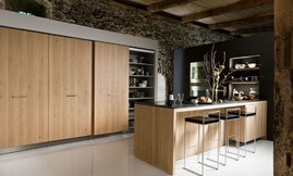 U-vormige keuken met keukeneiland en zitgelegenheid. Optisch wordt de keuken gedragen door het ruwe eiken fineer met verticale tekening. Een robuust oppervlakte contrasteert mooi met de koele rvs-apparatuur. Het keukeneiland is van een dun zwart glanzend aanrechtblad voorzien. Zuordnung: Stil Moderne keukens, Planungsart Keuken met keukeneiland