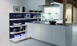 Deze open keuken heeft een gereduceerde look met witte fronten en een dun, metallic aanrechtblad. Het kookeiland staat haaks op een open wandgedeelte. Deze open wand met legplanken is ideaal om alles direct bij de hand te hebben, maar kan tegelijkertijd ook decoratief worden gebruikt. De blauwe achtergrond van de open kast zorgt voor een speels accent. Op deze open wandkast sluit het keukenblok met de spoelbak aan. De onder- en bovenkasten bieden voldoende bergruimte. Een keukenplanning zoals deze is ook erg geschikt voor grote ruimtes die deels als woonkamer functioneren. Zuordnung: Stil Design-keukens, Planungsart Binneninrichting van de keuken