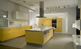 Eilandkeuken met fronten in de opvallende kleur mangogeel. De onderkasten van het keukenblok tegen de wand zijn geel, er zijn zowel lades als kasten. Het spatblad in de nis is wit, net als het aanrechtblad en de twee hangkasten boven de spoelbak. Het tweede keukenelement bestaat uit half hoge kasten waarin ook
de apparatuur is opgenomen. Ook deze kasten zijn allemaal mangogeel. Het kookeiland, eveneens mangogeel, heeft als fris accent ook een wit werkblad. Aan de zijkant steekt het werkbald iets uit waardoor er een zitgelegenheid voor twee personen is ontstaan. Onder het werkblad is er in het kookeiland nog eens extra bergruimte in twee normale kasten, twee lage kastjes en twee open kasten. Zuordnung: Stil Moderne keukens, Planungsart keukenblok