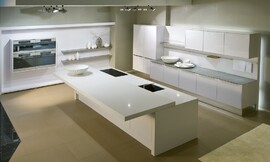 Een wit en rechthoekig kookeiland met uitstekend aanrecht om aan te zitten is het centrum van de keuken. De keukenwand heeft onder- en hoogkasten met fronten met greeplijsten en een dun aanrechtblad. Bij de andere muur ziet u hoe apparatuur in de muur ingebouwd kan worden. Zuordnung: Stil Moderne keukens, Planungsart Keuken met keukeneiland