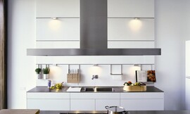 Designkeuken met symmetrische witte paneelwand waar overzichtelijk handige dingen als een kookboekstandaard, kruidenpotjes, messenrek of keukenpapierhouder aan kunnen worden gehangen. Bij deze keuken loopt de wand door tot aan het plafond. Vrij hoog zijn er nog twee spotjes geplaatst, maar ook op de hoogte van de kookboekhouder etc. zijn er kleine spotjes opgenomen. Dit zorgt voor een goede verlichting op het aanrechtblad en de dubbele spoelbak daaronder. De onderkastjes zijn greeploos en wit. Het aanrechtblad is dun en heeft een donkere kleur. Het kookeiland daar tegenover biedt ook naast het gasfornuis genoeg plaats voor het snijden, hakken en mengen van interessante recepten. Zuordnung: Stil Design-keukens, Planungsart Binneninrichting van de keuken