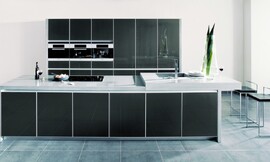 Elegantie en rechtlijnigheid in een donkere metallic tint achter glas met net aluminium kader. Hier ziet u ook het bijzondere effect van een aanrechtblad met verschillende hoogten. Rechts ziet u het verhoogde deel met de spoelbak en de eetbar. Zuordnung: Stil Design-keukens, Planungsart Open keuken (woonkeuken)